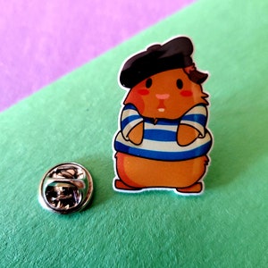 May include: A cartoon enamel pin of a brown and orange guinea pig wearing a blue and white striped shirt and a black beret. The pin has a silver backing.