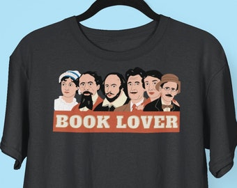 May include: A black t-shirt featuring a graphic of well-known authors, including Jane Austen, Charles Dickens, William Shakespeare, and Edgar Allan Poe. The text "BOOK LOVER" is printed in red and white on a red banner.