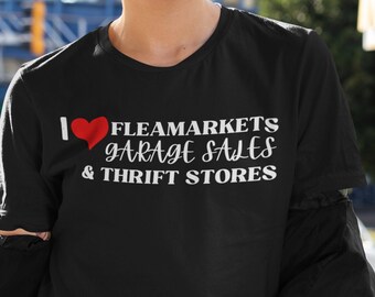 Puede incluir: Una camiseta negra con un gráfico blanco que dice "Me encantan los mercados de pulgas, las ventas de garaje y las tiendas de segunda mano". El gráfico incluye un corazón rojo.