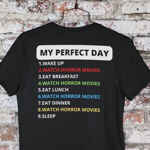 May include: Black t-shirt with white text that reads "My Perfect Day" and a list of activities, including "Watch Horror Movies" repeated multiple times.