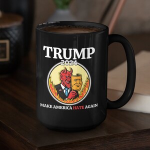 May include: Black ceramic mug featuring the text "TRUMP 2024" above an illustration of a devil and a mask. The phrase "MAKE AMERICA HATE AGAIN" is printed below the illustration. The mug is on a dark wooden surface.