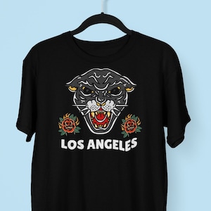 以下が含まれることがあります： 黒いTシャツに、黒い豹と赤いバラのグラフィックと、白い文字で「LOS ANGELES」とプリントされています。