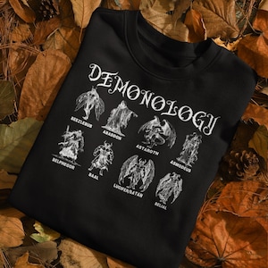 Könnte beinhalten: Schwarzes Sweatshirt mit dem Wort "Demonology" in gotischer Schrift, mit Illustrationen von Dämonen wie Beelzebub, Abaddon und Luzifer/Satan. Das Design ist in Weiß gedruckt.