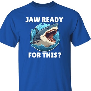 以下が含まれることがあります： ロイヤルブルーのTシャツに、口を開けたサメの白いグラフィックと「JAW READY FOR THIS?」のテキストがプリントされています