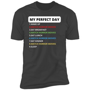 May include: A dark grey t-shirt with the text "MY PERFECT DAY" in white. The text is followed by a list of activities, including "WATCH HORROR MOVIES" in different colours.