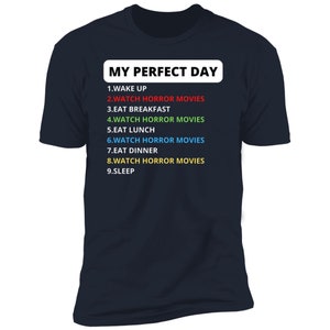 May include: A dark blue t-shirt with white text that reads "MY PERFECT DAY" and a list of activities, including "WATCH HORROR MOVIES" in different colours.