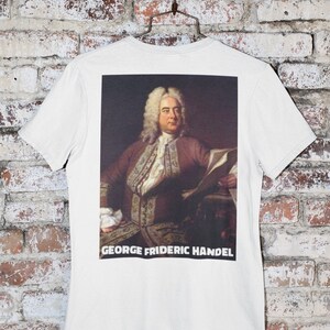 May include: White t-shirt with a printed image of George Frideric Handel, a famous composer, on the back. The text "GEORGE FRIDERIC HANDEL" is printed below the image.
