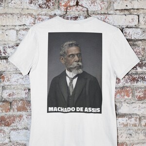 May include: White t-shirt with a black and white photo of Machado de Assis on the back. The photo shows a man in a suit and tie, with a beard and mustache.