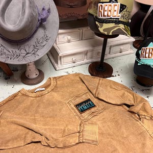 May include: A brown long-sleeved shirt with a distressed look and a blue patch that says "Fierce Female" in a pocket. The shirt is laid out on a white surface with two hats and a small white dresser in the background.