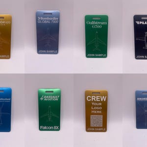 May include: Eight different metal luggage tags with various aircraft models and company logos. The tags are all silver, gold, blue, or green. The text on the tags includes "Praetor 600", "Bombardier Global 7500", "Gulfstream G700", "Pilatus", "AgustaWestland", "Dassault Aviation Falcon 8X", "Crew Your Logo Here", and "Sikorsky".