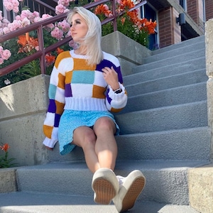 Puede incluir: Una mujer con un suéter de patchwork blanco, morado, naranja y azul, una falda azul claro y zapatillas blancas. Está sentada en un tramo de escaleras de hormigón.