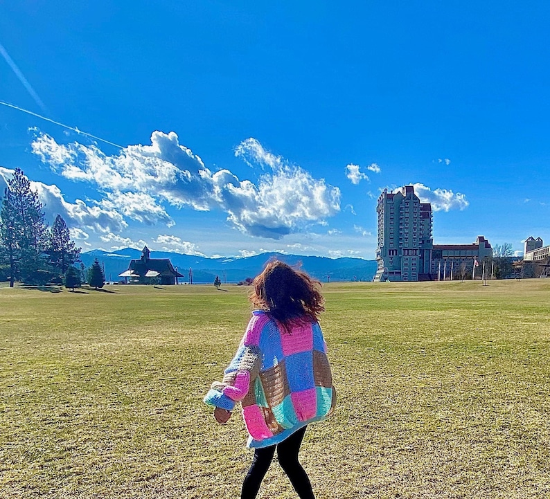 Pu&ograve; includere: Una persona che indossa un cardigan patchwork colorato in un campo con un edificio sullo sfondo.