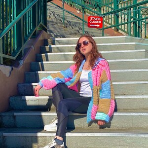 Pu&ograve; includere: Una donna indossa un cardigan all'uncinetto colorato, leggings neri e scarpe da ginnastica bianche e siede su una scalinata. Un cartello rosso con la scritta "STAIRS CLOSED" e una freccia che punta a destra &egrave; visibile sullo sfondo.