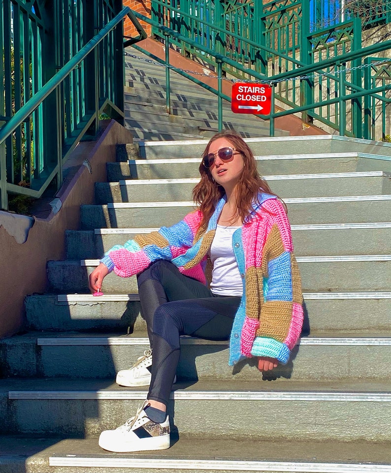 Pu&ograve; includere: Una donna indossa un cardigan all'uncinetto colorato, leggings neri e scarpe da ginnastica bianche e siede su una scalinata. Un cartello rosso con la scritta "STAIRS CLOSED" e una freccia che punta a destra &egrave; visibile sullo sfondo.