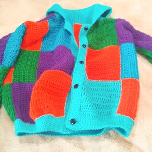 Pu&ograve; includere: Un cardigan all'uncinetto colorato con cappuccio e chiusura a bottoni. Il cardigan &egrave; realizzato in stile patchwork con quadrati di diversi colori, tra cui verde, viola, arancione e turchese.