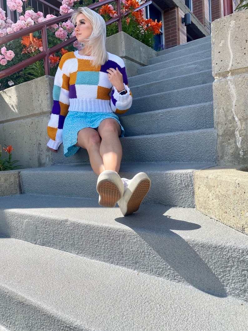 May include: A woman wearing a white, purple, orange and blue patchwork sweater with a light blue skirt and white trainers. She is sitting on a set of concrete steps.