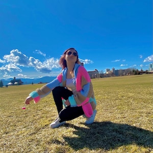 Pu&ograve; includere: Una donna indossa un cardigan all'uncinetto colorato con una canotta bianca e leggings neri. &Egrave; accovacciata in un campo erboso con un cielo blu sullo sfondo.