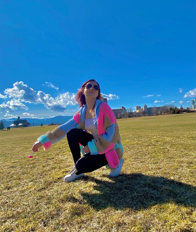 Pu&ograve; includere: Una donna indossa un cardigan all'uncinetto colorato con una canotta bianca e leggings neri. &Egrave; accovacciata in un campo erboso con un cielo blu sullo sfondo.