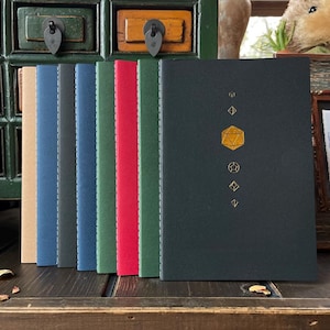 May include: A selection of colourful notebooks, one with a dark green cover and gold geometric design. The notebooks are in shades of tan, blue, green, and red. They are displayed on a dark wooden surface, likely a table.