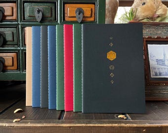 May include: A collection of colorful notebooks with a dark green cover featuring a gold geometric design. The notebooks are in various colors, including tan, blue, green, and red. The notebooks are displayed on a dark wooden surface.
