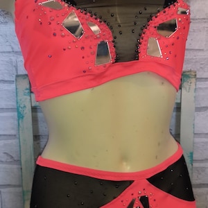 Op de afbeelding: Een tweedelig danskostuum in koraalroze en zwart. De top heeft een halternek en mesh details, versierd met spiegelaccenten en strass. De bijpassende broek heeft een vergelijkbaar ontwerp met een geometrisch patroon en extra versieringen.