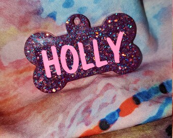 May include: A pink glitter dog tag shaped like a bone with the name "HOLLY" written in pink letters.