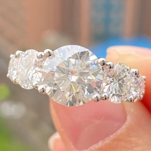 Könnte beinhalten: Ein silberner Ring mit fünf runden, klaren Edelsteinen. Der Mittelstein ist der größte, flankiert von zwei kleineren Steinen auf jeder Seite. Der Ring wird vor einem verschwommenen Hintergrund gehalten.