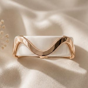 May include: A ring featuring a smooth white band accented by a polished rose gold-toned metal overlay that forms a continuous wave pattern across the center. The design is symmetrical, with the metallic wave rising and dipping along the white surface.