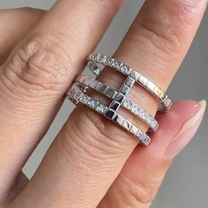 May include: A silver-toned multi-band ring featuring three parallel horizontal bars connected by a vertical bridge. Each bar is embedded with a series of small, clear, square-cut stones that span the entire length of the metal bands.