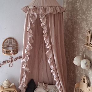 以下が含まれることがあります： 子供部屋に、フリル付きの縁が付いたピンク色の天蓋が吊り下げられています。白いマットレス、ぬいぐるみ、キノコ型のスツールが床にあります。ラタンの棚と装飾的なキャビネットも表示されます。