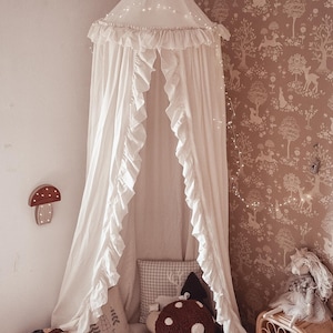 May include: A white canopy bed with ruffled edges and fairy lights hangs from the ceiling. The bed is decorated with pillows and a mushroom-shaped cushion. A mushroom-shaped wall light and a woven basket are also visible.
