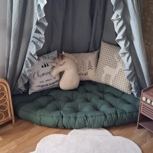 Può includere: Un'area giochi per bambini con un baldacchino con bordi a balze, un cuscino verde trapuntato e cuscini decorativi. Un tappeto bianco a forma di nuvola è sul pavimento di legno. I cuscini hanno disegni natalizi e a tema foresta.