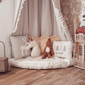 Puede incluir: Un rincón de lectura para niños con un dosel crema y ribete con volantes. El espacio incluye un colchón blanco, cojines decorativos con un diseño de ciervo, unicornio y bosque, y un oso de peluche. También se ve una casa de muñecas de madera.