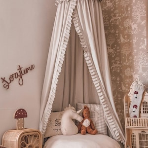 Può includere: Una scena di asilo nido dai toni neutri con un letto a baldacchino con finiture a balze, un cuscino rotondo e cuscini decorativi. Un comodino in legno con una lampada a fungo e una casa delle bambole aggiungono all'arredamento stravagante. Il muro presenta un cartello "Storytime".