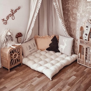 May include: A cozy reading nook with a cream-colored canopy and a floor cushion. Decorative pillows in various patterns and colors, including a tree-shaped pillow, add to the inviting atmosphere. A wooden dollhouse and a rattan side table complete the scene.