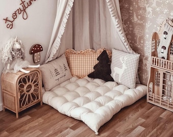 May include: A cozy reading nook with a cream-colored canopy and a floor cushion. Decorative pillows in various patterns and colors, including a tree-shaped pillow, add to the inviting atmosphere. A wooden dollhouse and a rattan side table complete the scene.