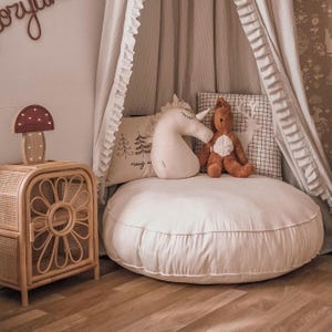 May include: A cozy nursery scene featuring a rattan nightstand with a mushroom lamp, a white floor cushion, and a canopy. Decorative pillows include a unicorn and a deer. The room has a soft, neutral color palette.