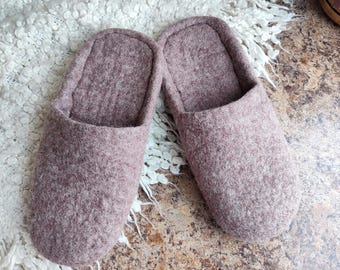 May include: A pair of mauve-colored felted wool slippers. The slippers have a simple, slip-on design with a rounded toe and a soft, textured appearance. They are resting on a white, textured surface.