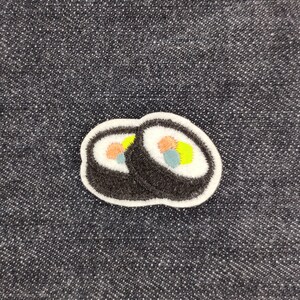 Op de afbeelding: Twee geborduurde sushi-rollen met witte contouren en kleurrijke vullingen op een donkerblauwe denim achtergrond.