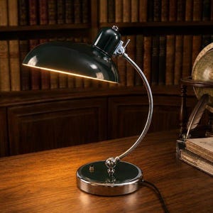Op de afbeelding: Een donkergroene bureaulamp met een gebogen chromen arm en een ronde chromen voet. De lamp heeft een brede, koepelvormige kap die het licht naar beneden richt, en een kleine drukknopschakelaar bevindt zich bovenop de ronde voet.