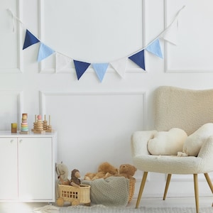 Könnte beinhalten: Ein weißer Schrank mit zwei Türen und ein weißer Sessel mit einem weißen Kissen. Vor dem Sessel steht ein Korb mit Spielzeug. In der Ecke des Raumes steht eine Stehlampe aus Holz mit einem weißen Lampenschirm. Über dem Schrank hängt eine blau-weiße Girlande.