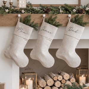 Könnte beinhalten: Drei weihnachtliche Strümpfe aus beigem Stoff hängen an einem Kaminsims aus Holz. Jeder Strumpf hat ein Bündchen mit eingesticktem schwarzem Text, der Mom, James und Dad lautet. Grüne Tannenzweige stecken in jedem der Strümpfe.