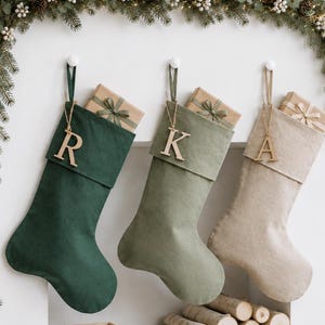 May include: Three Christmas stockings in dark green, sage green, and beige, each featuring a wooden letter tag R, K, or A. Each stocking contains a small gift box wrapped in brown paper with a green ribbon. A wooden block calendar shows December 25.