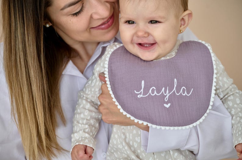 May include: A dusty purple baby bib featuring a white pom-pom trim along the edge. The centre of the bib is embroidered with the name Layla in white cursive script, positioned above a small white heart icon, creating a simple and clean design.