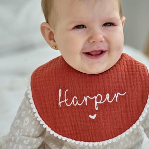 May include: A round, rust-coloured muslin bib featuring the embroidered white name Harper in a cursive script, accented with a small white heart below the name and a delicate white pom-pom trim along the entire outer edge of the fabric.