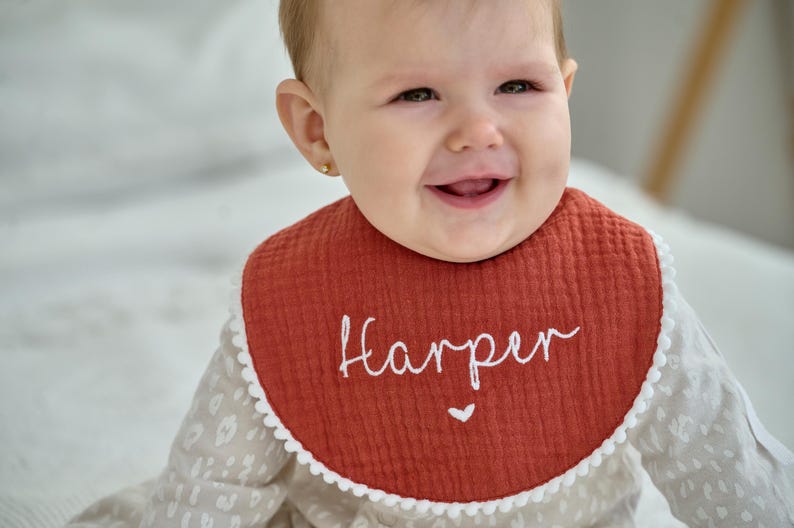 May include: A round, rust-coloured muslin bib featuring the embroidered white name Harper in a cursive script, accented with a small white heart below the name and a delicate white pom-pom trim along the entire outer edge of the fabric.