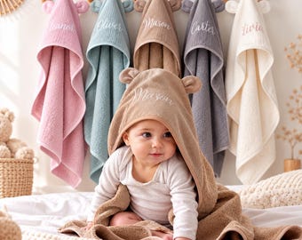 Peut inclure: Une collection de cinq serviettes à capuche en rose, bleu, beige, gris et blanc, chacune avec des oreilles d'ours sur la capuche et des noms brodés. Une serviette beige est drapée sur un enfant portant un body blanc à manches longues.