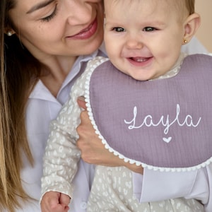 May include: A woman holding a baby wearing a purple and white bib with the name "Layla" embroidered on it.