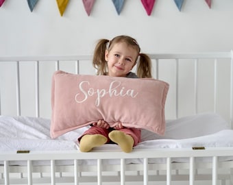 May include: A young girl sits in a white cot holding a pink pillow with the name "Sophia" embroidered in white.