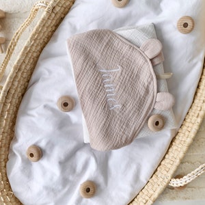 May include: A beige hooded baby towel with the name "James" embroidered on it. The towel is folded and placed in a wicker basket with wooden beads.